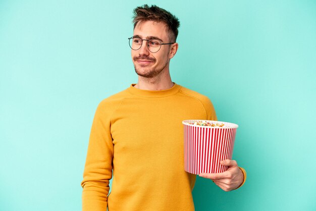 Young caucasian man eating popcorn isolated on blue background looks aside smiling, cheerful and pleasant.