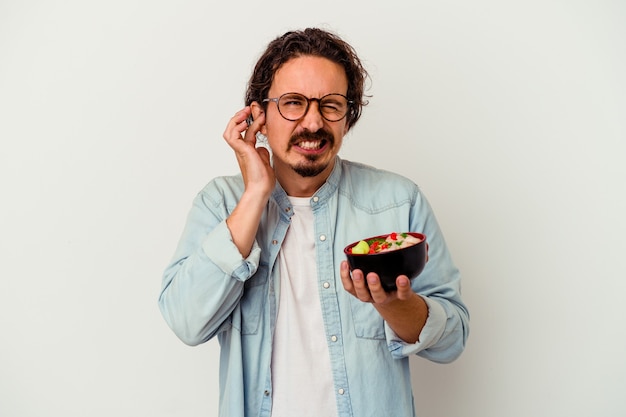 Young caucasian man eating a ramen isolated on white wall covering ears with hands