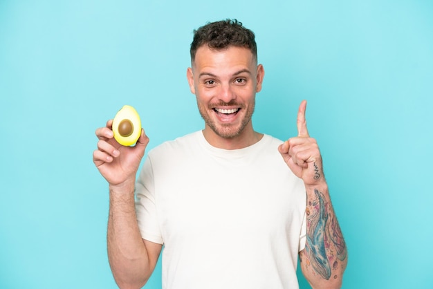 Young caucasian man holding an avocado isolated on blue background pointing up a great idea