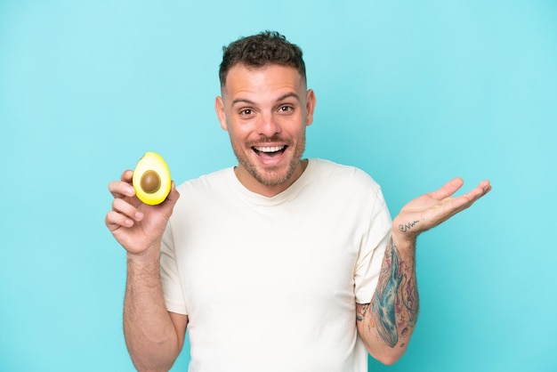 Young caucasian man holding an avocado isolated on blue background with shocked facial expression