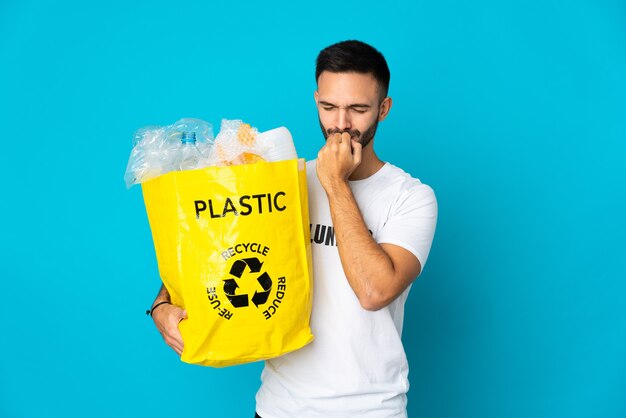 Young caucasian man holding a bag full of plastic bottles to recycle isolated on blue wall having doubts