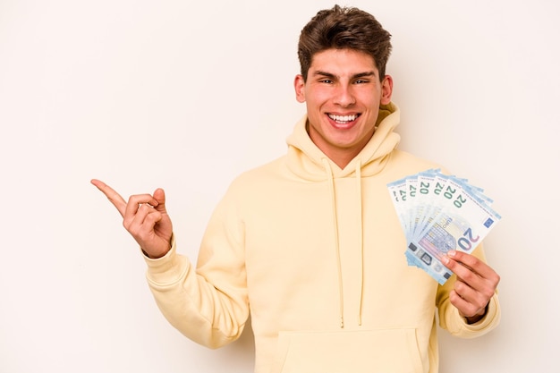 Young caucasian man holding banknotes isolated on white background smiling and pointing aside showing something at blank space