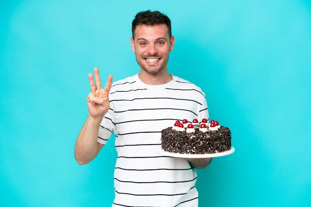 Young caucasian man holding birthday cake isolated on blue background happy and counting three with fingers