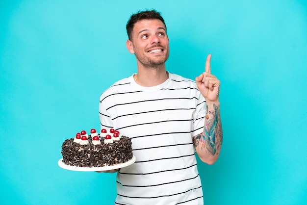 Young caucasian man holding birthday cake isolated on blue background pointing up a great idea