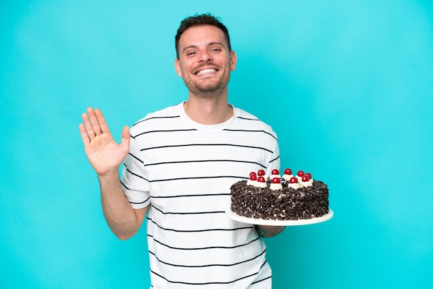 Young caucasian man holding birthday cake isolated on blue background saluting with hand with happy expression