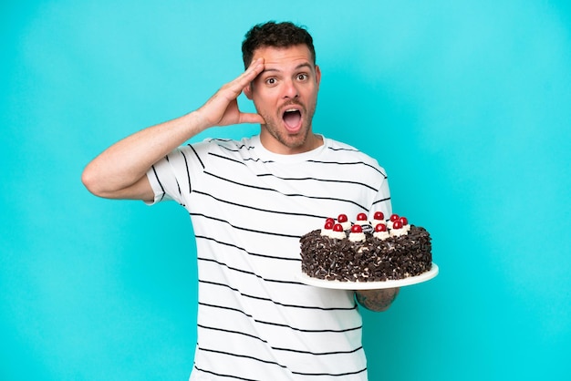 Young caucasian man holding birthday cake isolated on blue background with surprise expression