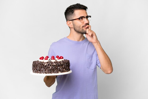 Young caucasian man holding birthday cake isolated on white background having doubts while looking up