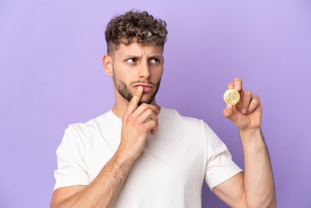 Young caucasian man holding a Bitcoin isolated on purple background having doubts while looking up