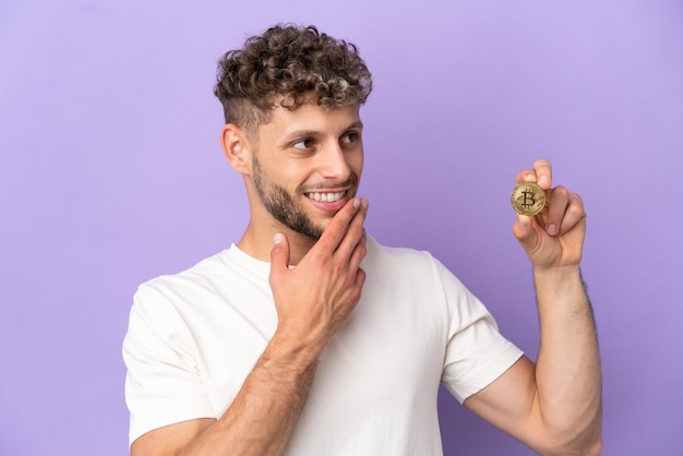 Young caucasian man holding a Bitcoin isolated on purple background looking up while smiling