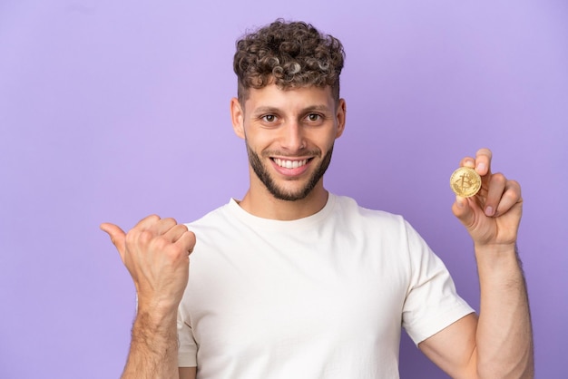 Young caucasian man holding a Bitcoin isolated on purple background pointing to the side to present a product