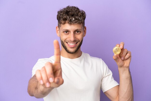 Young caucasian man holding a Bitcoin isolated on purple background showing and lifting a finger