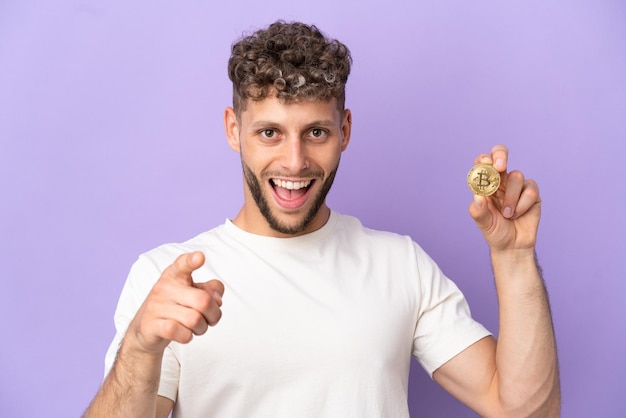 Young caucasian man holding a Bitcoin isolated on purple background surprised and pointing front