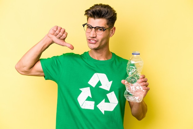 Young caucasian man holding a bottle of plastic to recycle isolated on yellow background feels proud and self confident, example to follow.