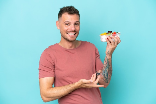 Young caucasian man holding a bowl of fruit isolated on blue background extending hands to the side for inviting to come