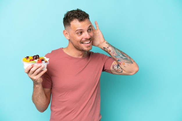 Young caucasian man holding a bowl of fruit isolated on blue background listening to something by putting hand on the ear