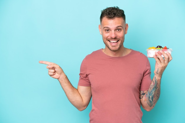 Young caucasian man holding a bowl of fruit isolated on blue background pointing finger to the side