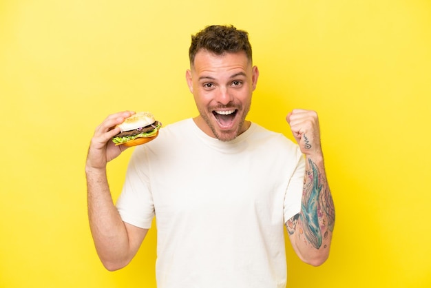 Young caucasian man holding a burger isolated on yellow background celebrating a victory in winner position