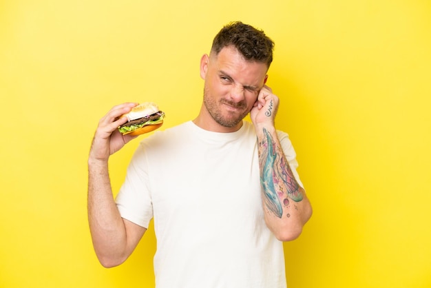 Young caucasian man holding a burger isolated on yellow background frustrated and covering ears