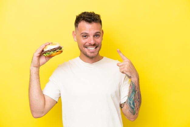 Young caucasian man holding a burger isolated on yellow background giving a thumbs up gesture