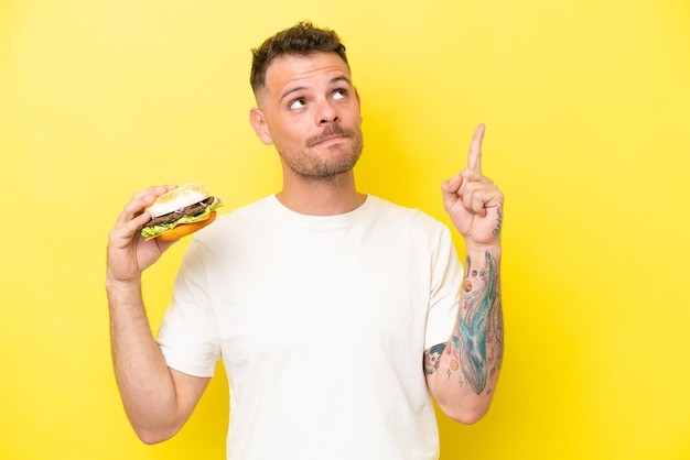 Young caucasian man holding a burger isolated on yellow background pointing up a great idea