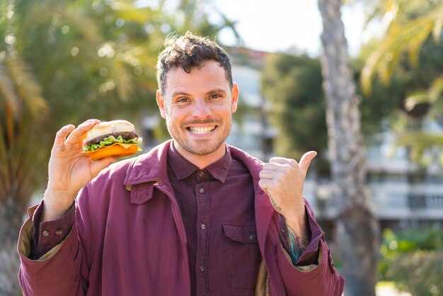 Young caucasian man holding a burger at outdoors pointing to the side to present a product