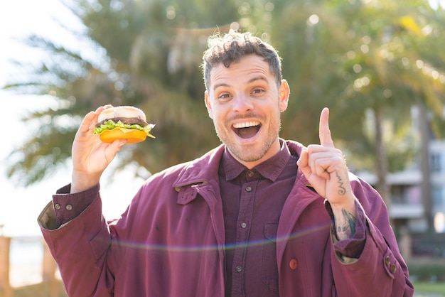 Young caucasian man holding a burger at outdoors pointing up a great idea