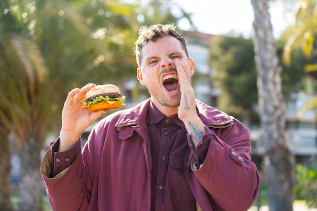 Young caucasian man holding a burger at outdoors shouting with mouth wide open