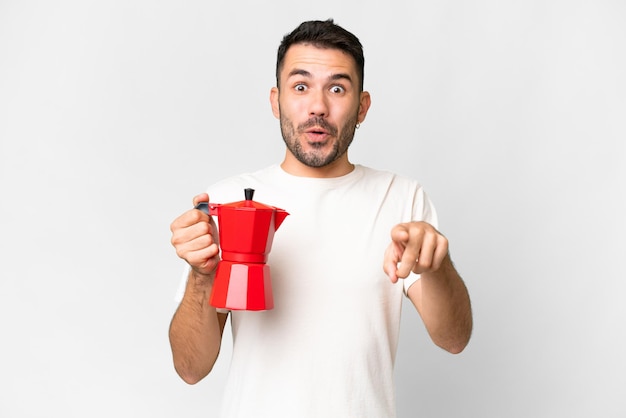 Young caucasian man holding coffee pot over isolated white background surprised and pointing front