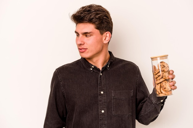 Young caucasian man holding cookies jar isolated on white background looks aside smiling cheerful and pleasant