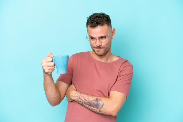 Young caucasian man holding cup of coffee isolated on blue background with sad expression