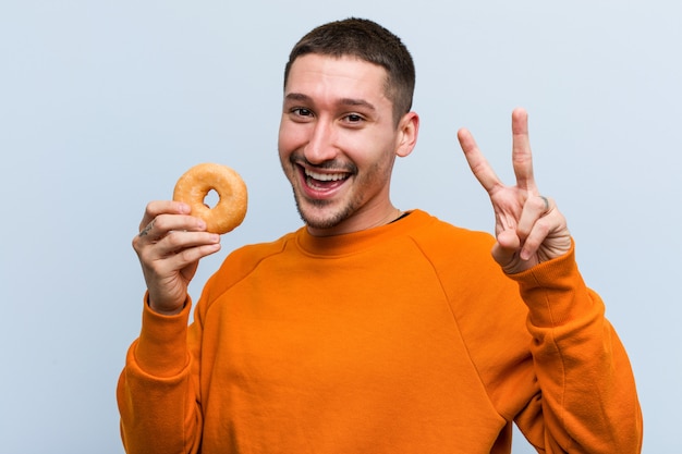 Young caucasian man holding a donut showing number two with fingers.