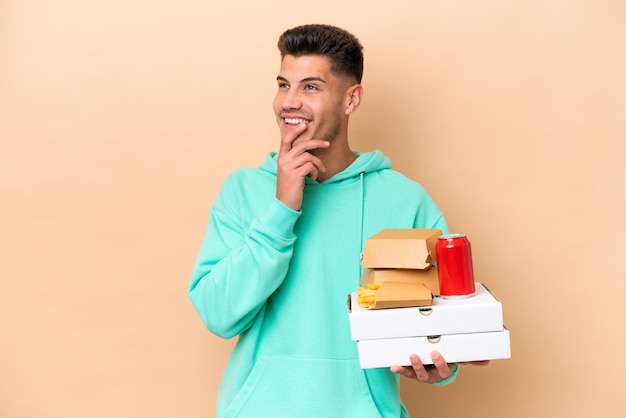 Young caucasian man holding fast food isolated on beige background looking up while smiling