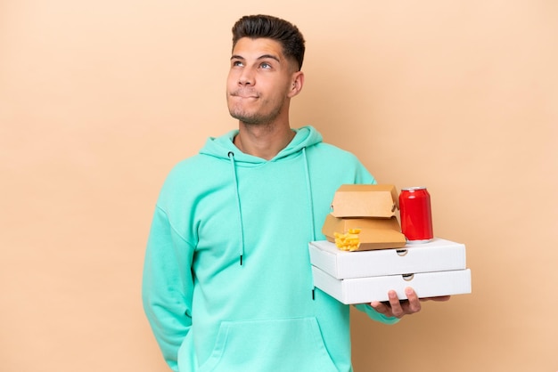 Young caucasian man holding fast food isolated on beige background and looking up