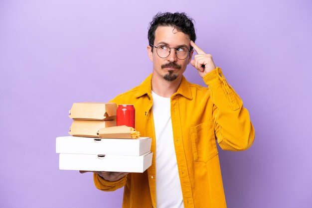 Young caucasian man holding fast food isolated on purple background having doubts and thinking