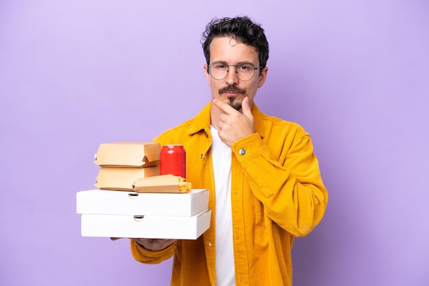 Young caucasian man holding fast food isolated on purple background thinking