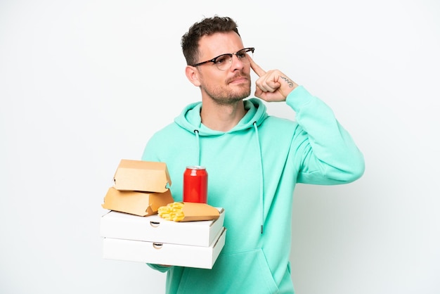 Young caucasian man holding fast food isolated on white background having doubts and thinking