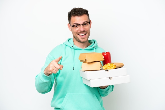 Young caucasian man holding fast food isolated on white background surprised and pointing front