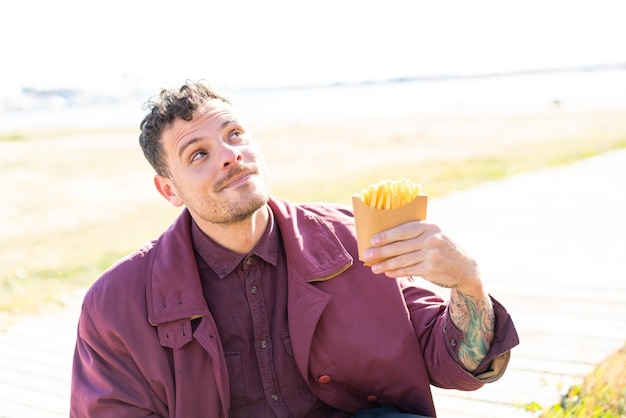 Young caucasian man holding fried chips at outdoors looking up while smiling