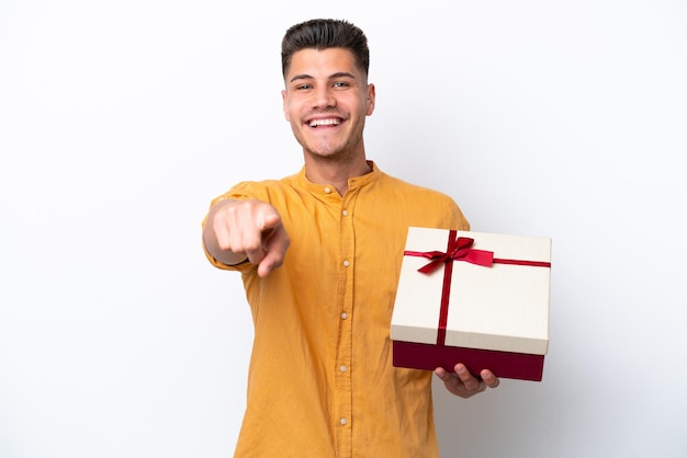 Young caucasian man holding a gift isolated on white background points finger at you with a confident expression