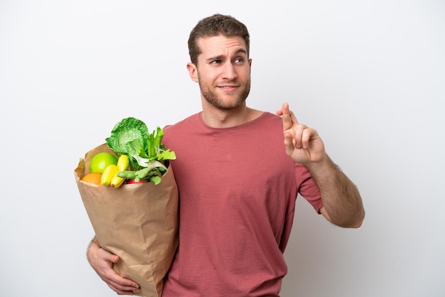 Young caucasian man holding a grocery shopping bag isolated on white background with fingers crossing and wishing the best