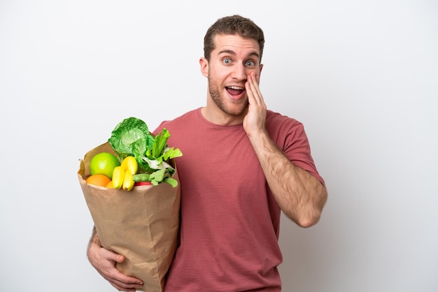 Young caucasian man holding a grocery shopping bag isolated on white background with surprise and shocked facial expression