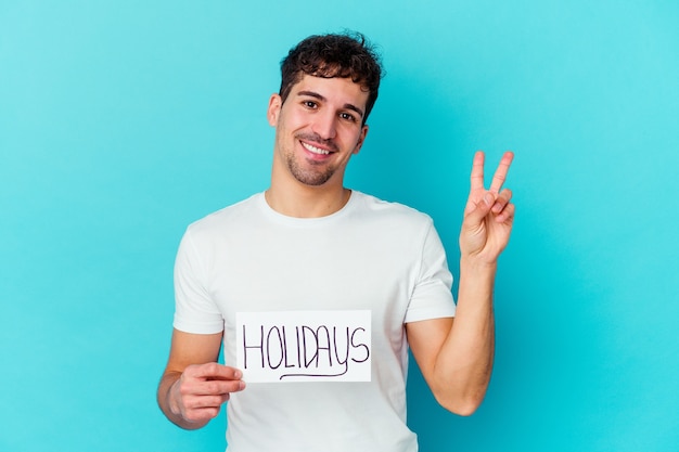 Young caucasian man holding a holidays placard joyful and carefree showing a peace symbol with fingers.