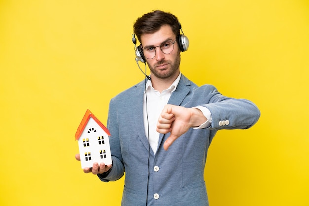 Young caucasian man holding a house isolated on white background showing thumb down with negative expression