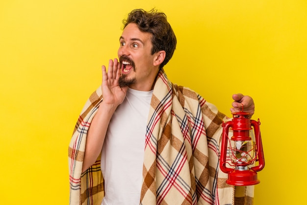 Young caucasian man holding lantern isolated on yellow background shouting and holding palm near opened mouth.