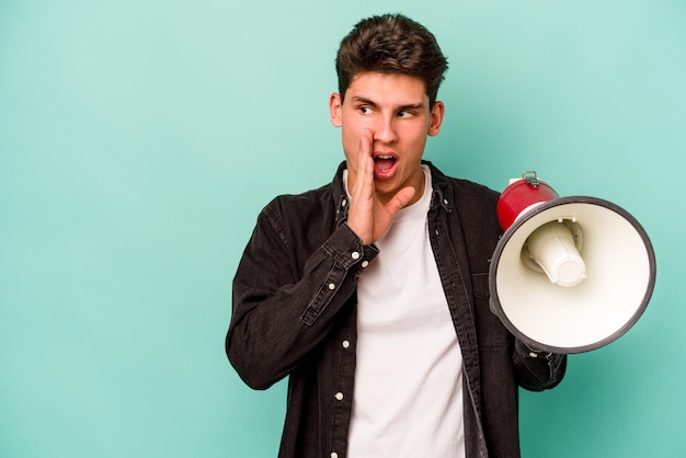 Young caucasian man holding a megaphone isolated on white background is saying a secret hot braking news and looking aside