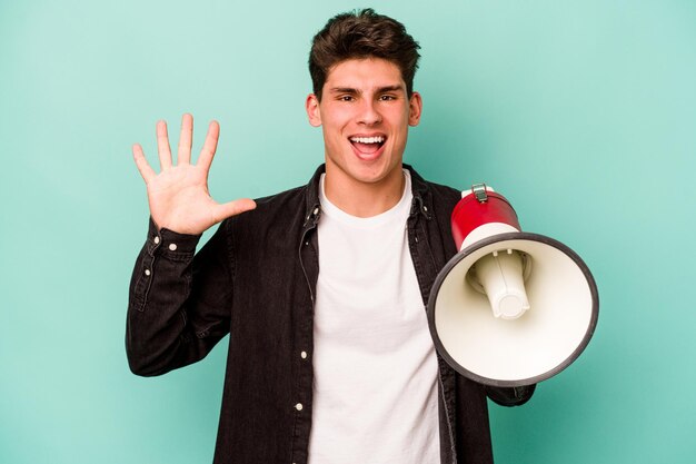 Young caucasian man holding a megaphone isolated on white background smiling cheerful showing number five with fingers