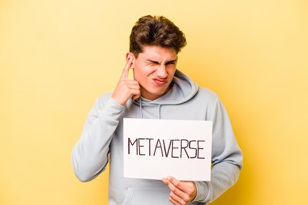 Young caucasian man holding metaverse placard isolated on yellow background covering ears with hands