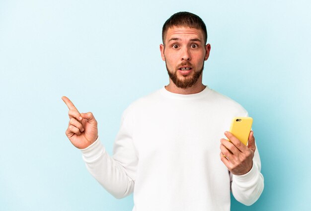 Young caucasian man holding mobile phone isolated on blue background pointing to the side