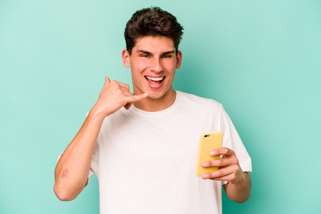 Young caucasian man holding mobile phone isolated on blue background showing a mobile phone call gesture with fingers