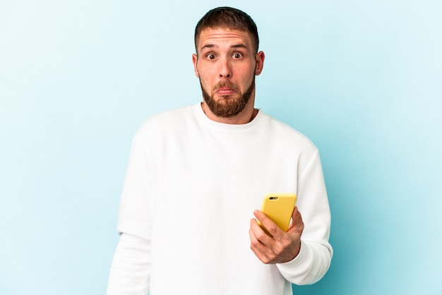 Young caucasian man holding mobile phone isolated on blue background shrugs shoulders and open eyes confused.